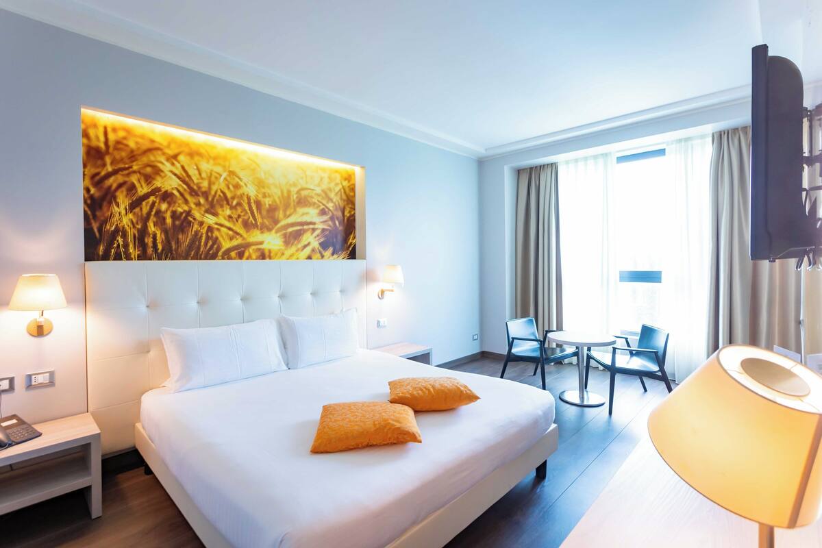 Une chambre d’hôtel avec un grand lit, deux oreillers, une table de chevet avec une lampe, une télévision, un bureau avec une chaise et une fenêtre avec des rideaux.