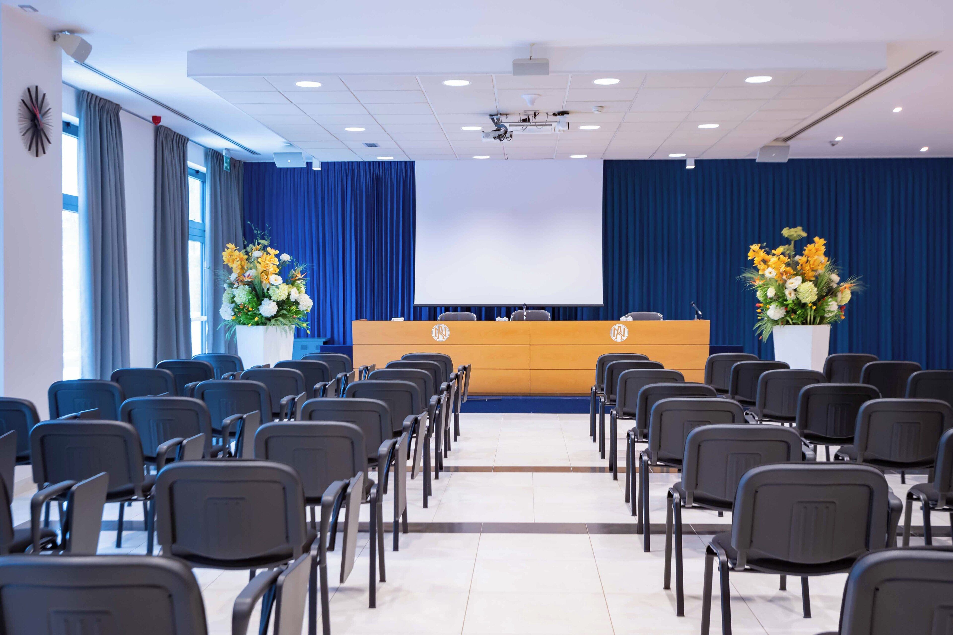 A conference room with rows of chairs, a podium, and two flower arrangements.