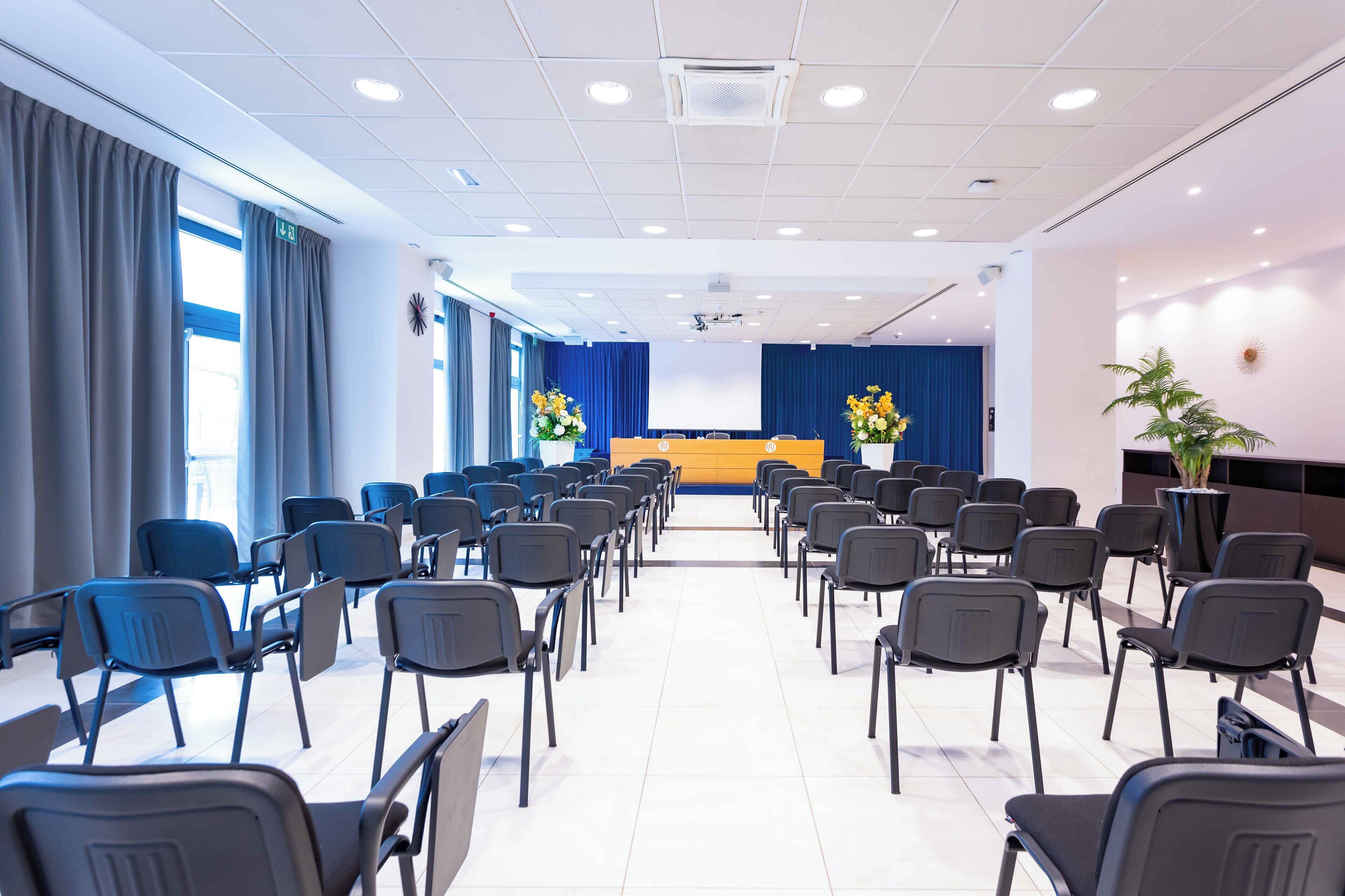 Un salón de conferencias con filas de sillas, un atril y una pantalla grande.