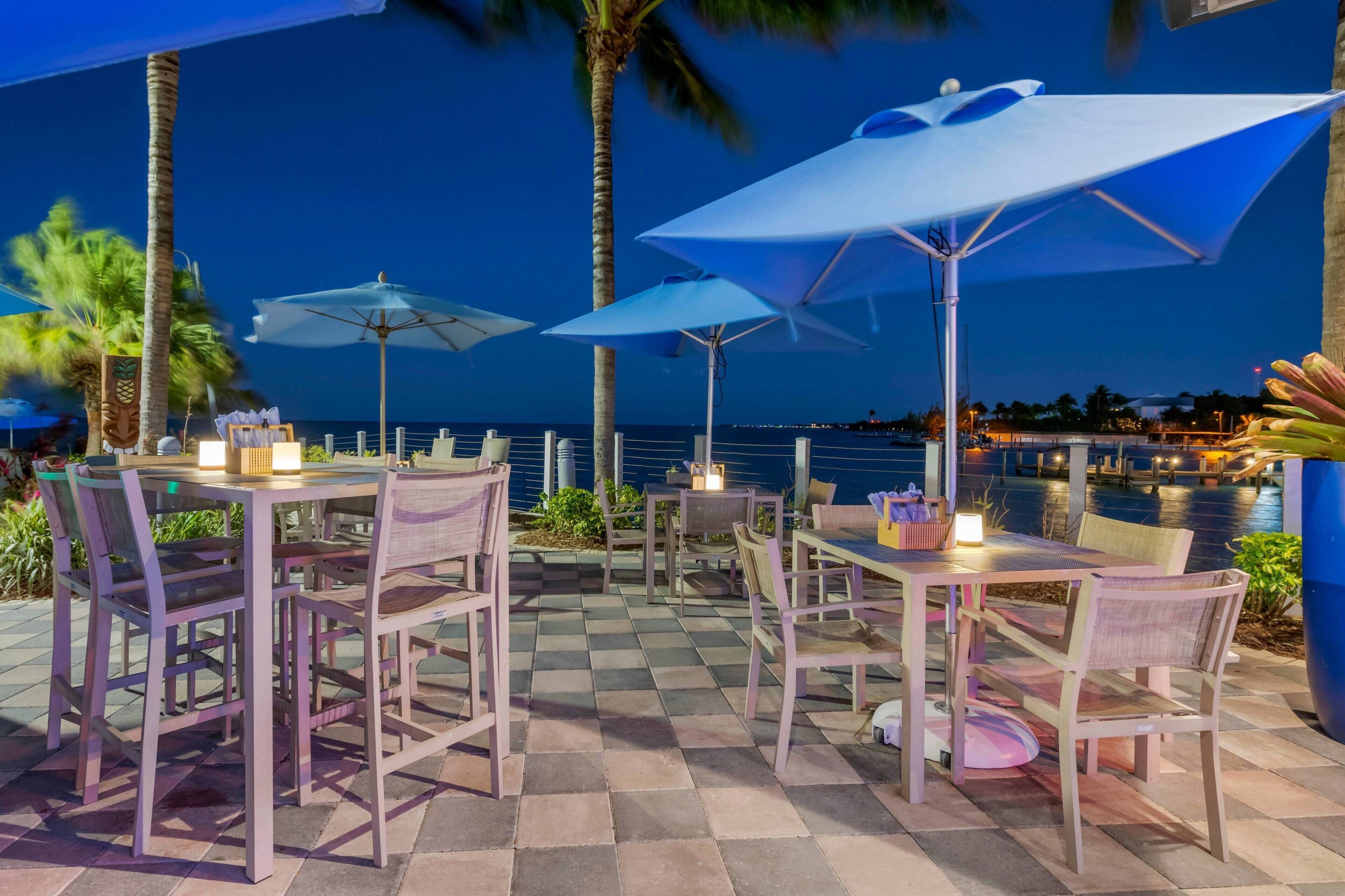 Un espace extérieur aménagé pour les repas, avec des tables, des chaises, des parasols et une vue sur l’océan au crépuscule.