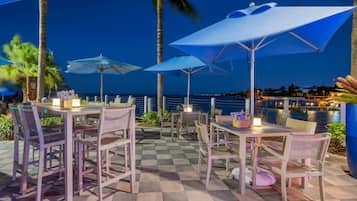 Un espace extérieur aménagé pour les repas, avec des tables, des chaises, des parasols et une vue sur l’océan au crépuscule.