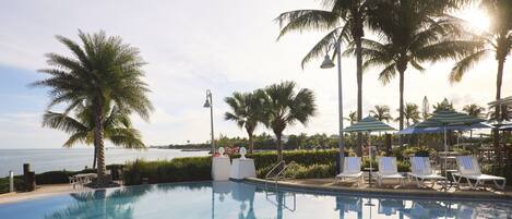 A pool area with lounge chairs and umbrellas, surrounded by palm trees and a view of the ocean.