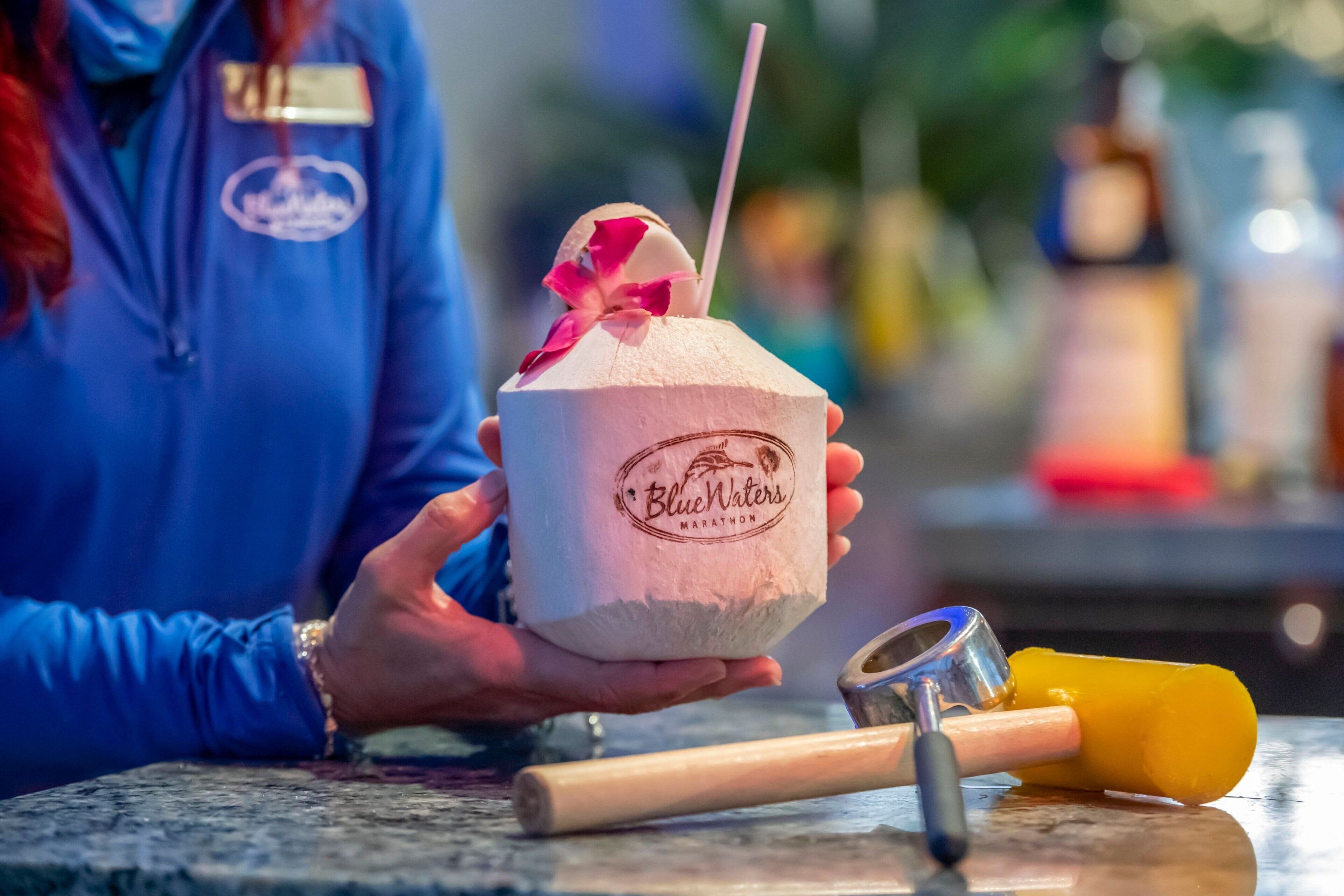 Una persona sostiene un vaso de papel con pajita y una flor, con el texto “Blue Waters Marathon”. Hay una paleta de hielo amarilla y un recipiente metálico sobre una encimera.