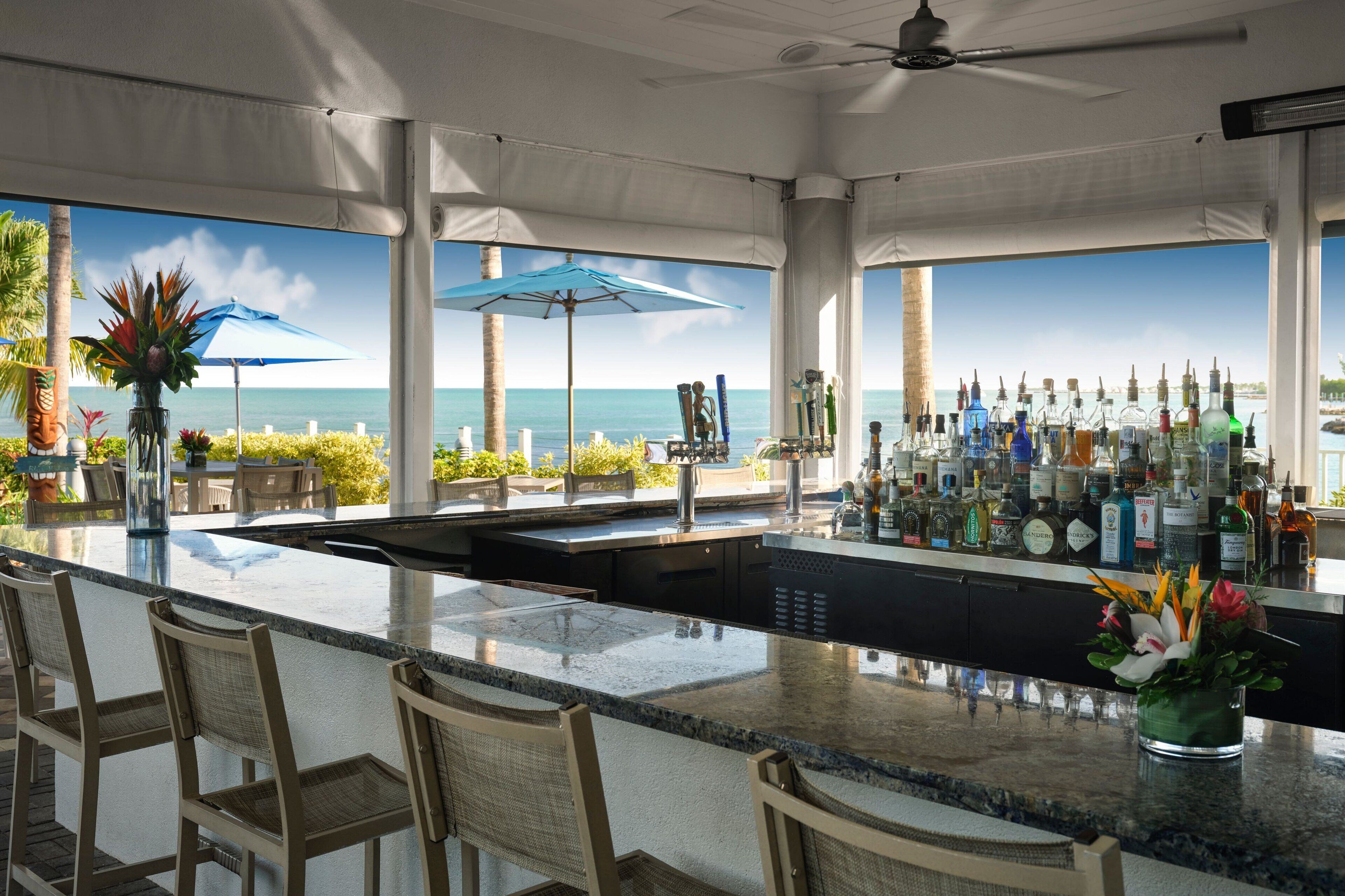 A bar area with a view of the ocean, bar stools, and a variety of bottles.