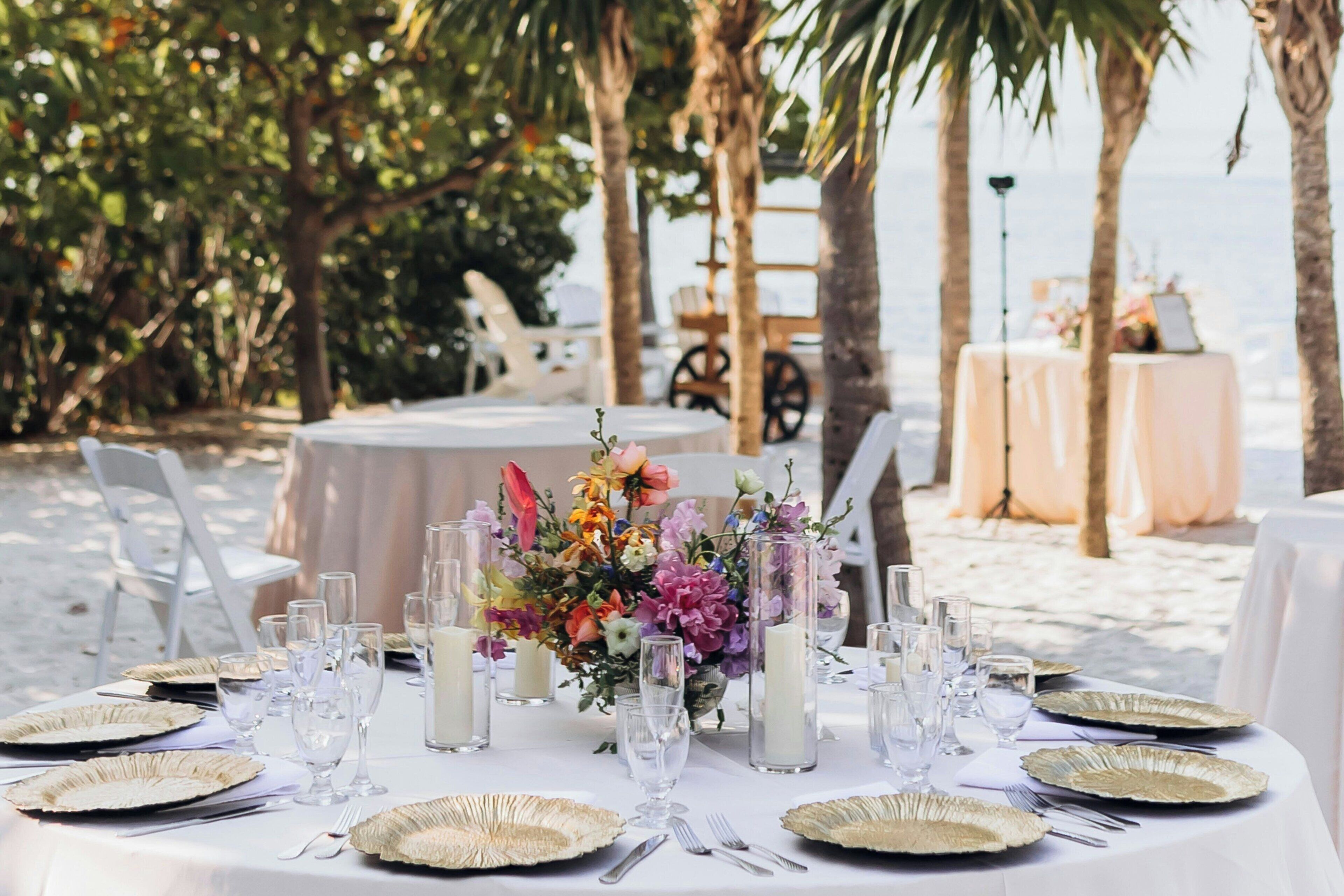 A beachside dining setup with round tables, white tablecloths, and gold-rimmed plates.
