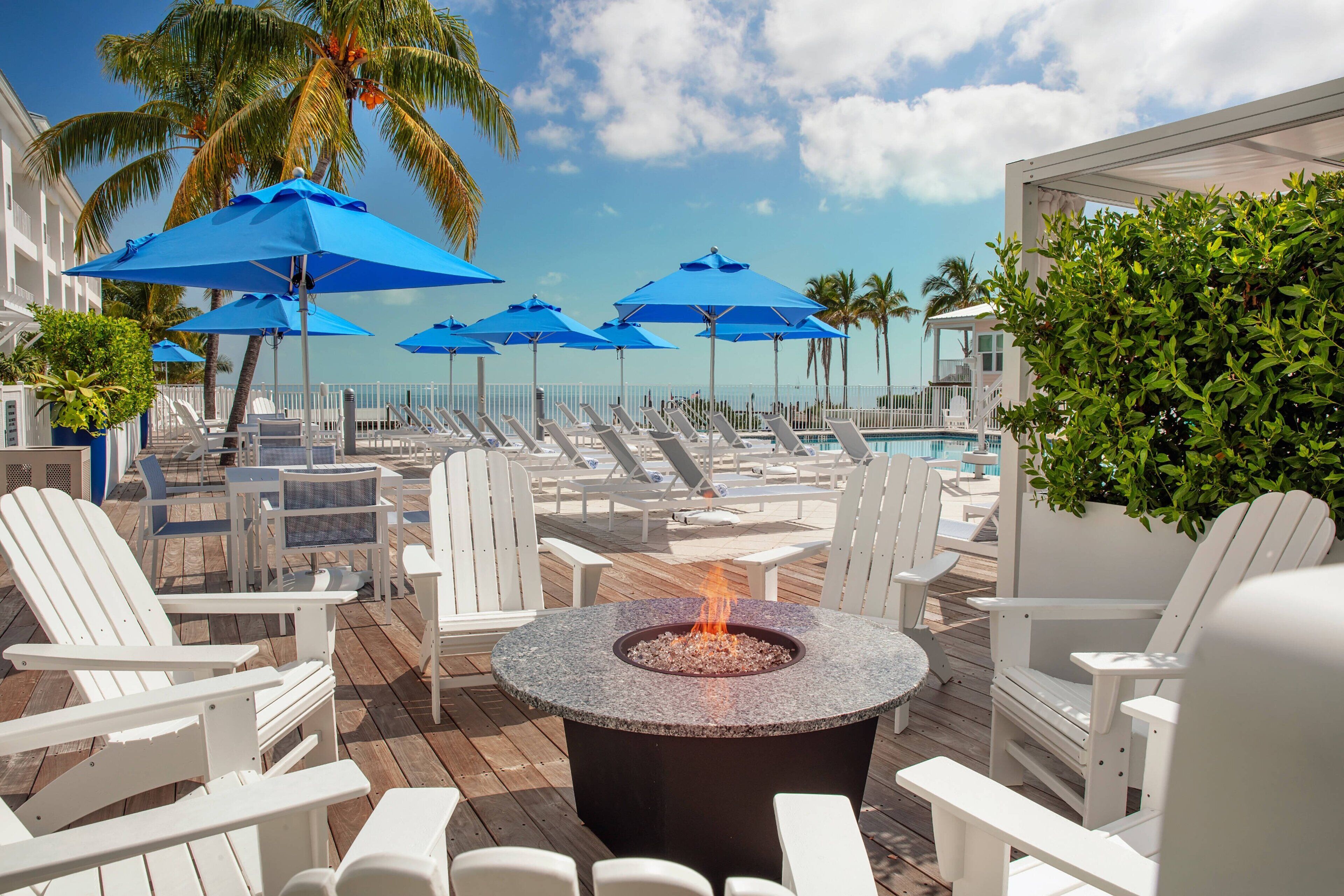 A poolside area with lounge chairs, blue umbrellas, and a fire pit.