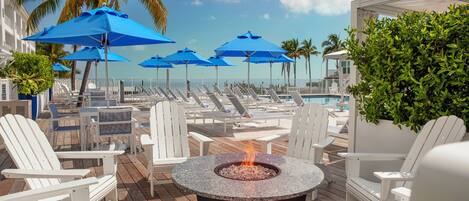 A poolside area with lounge chairs, blue umbrellas, and a fire pit.