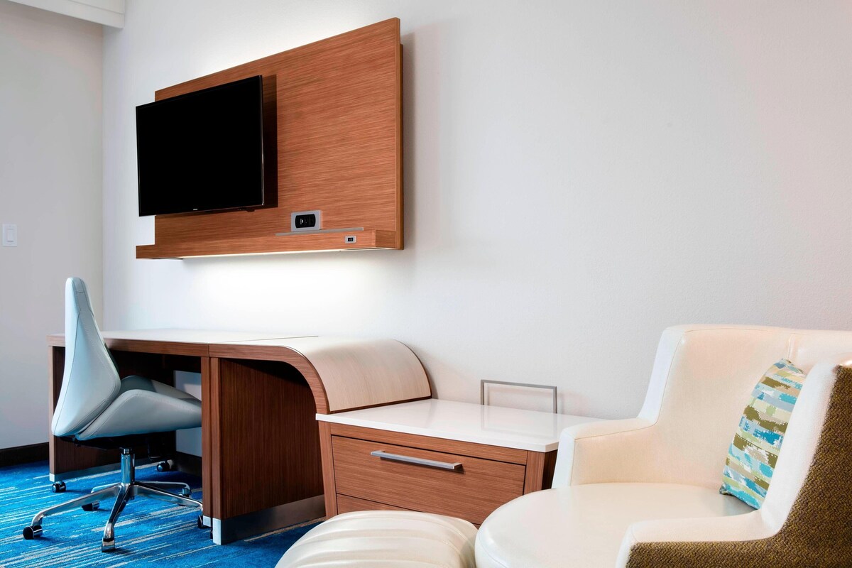 A modern hotel room with a flat-screen TV mounted on the wall, a desk with a chair, a sofa, and a small bedside table.
