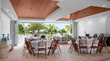 Uma área de refeições ao ar livre com mesas e cadeiras de madeira, um buffet e vista para palmeiras e o oceano.