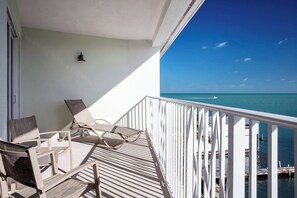 A balcony with two chairs and a table, overlooking a clear blue sea and a dock with boats.
