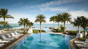 A clear blue pool with white lounge chairs and palm trees, overlooking a calm sea.
