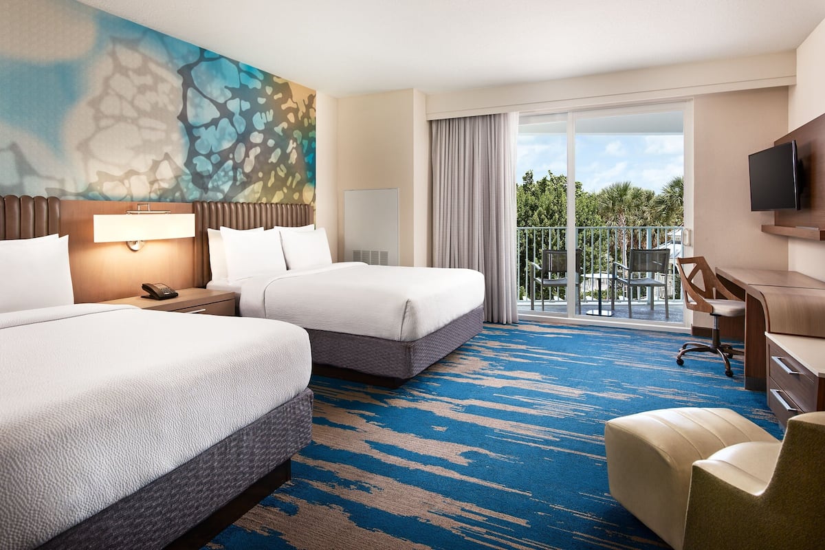 A hotel room with two beds, a desk, a chair, a TV, and a balcony with a view of palm trees.