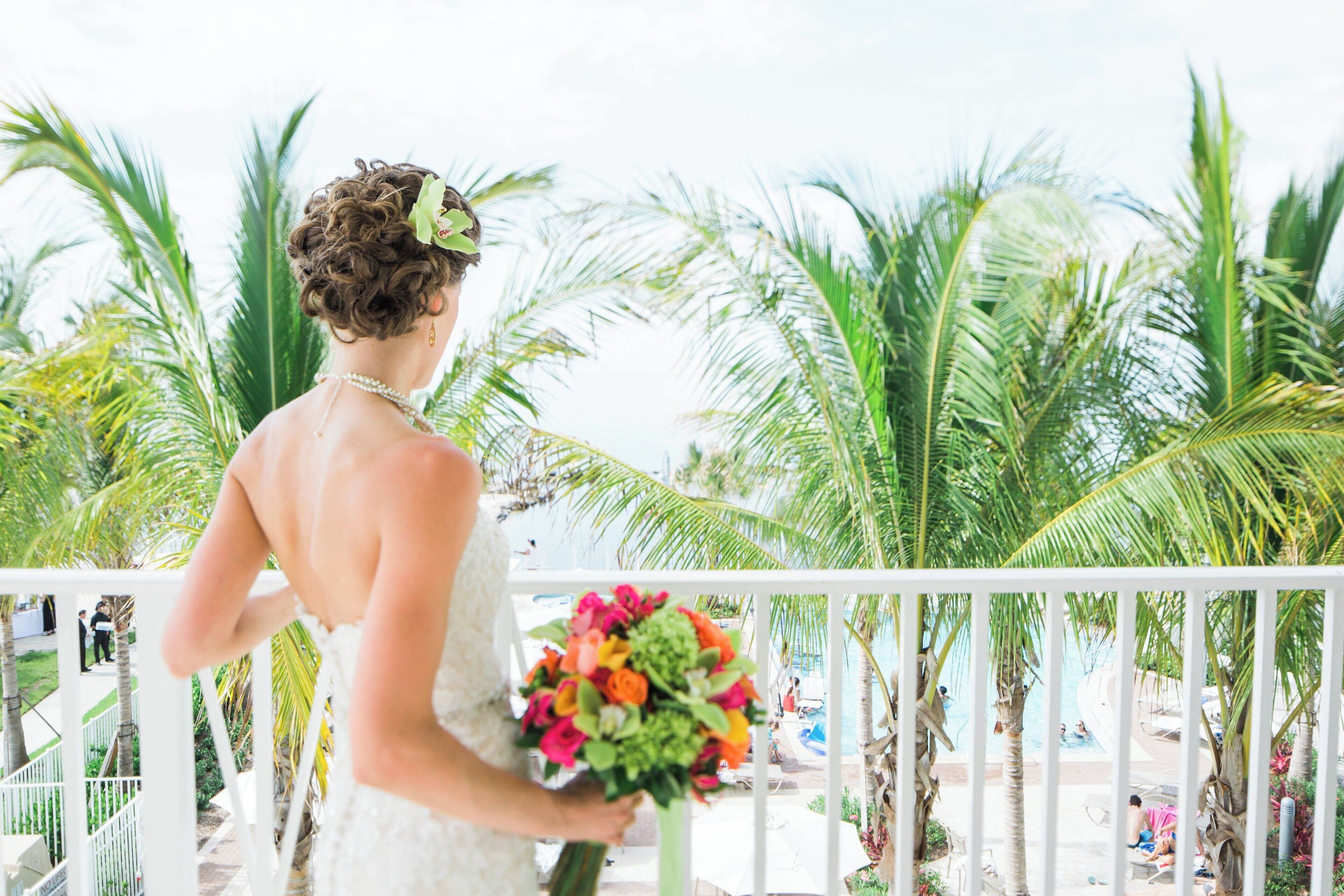 A bride holding a bouquet on a balcony with a view of palm trees and a pool.