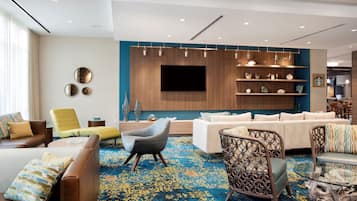 A modern hotel lobby with a large seating area, a flat-screen TV mounted on the wall, and a Starbucks coffee machine.
