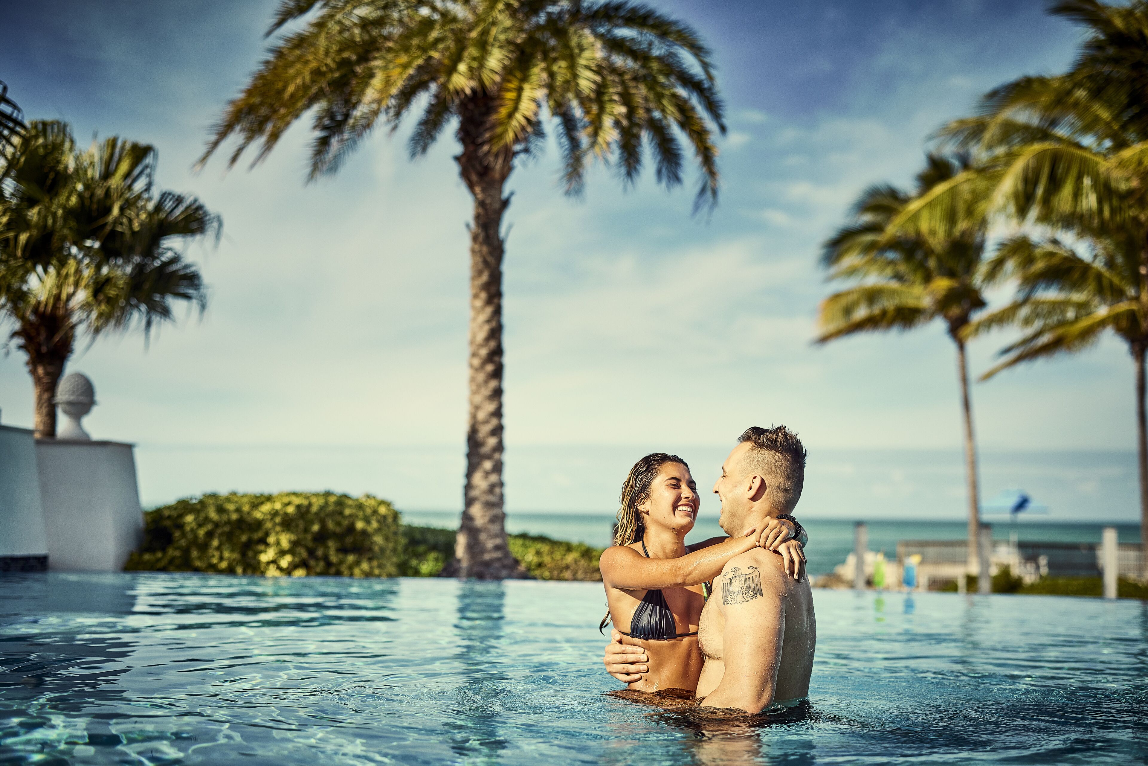 A couple embracing in a clear blue infinity pool with palm trees and a view of the ocean in the background.