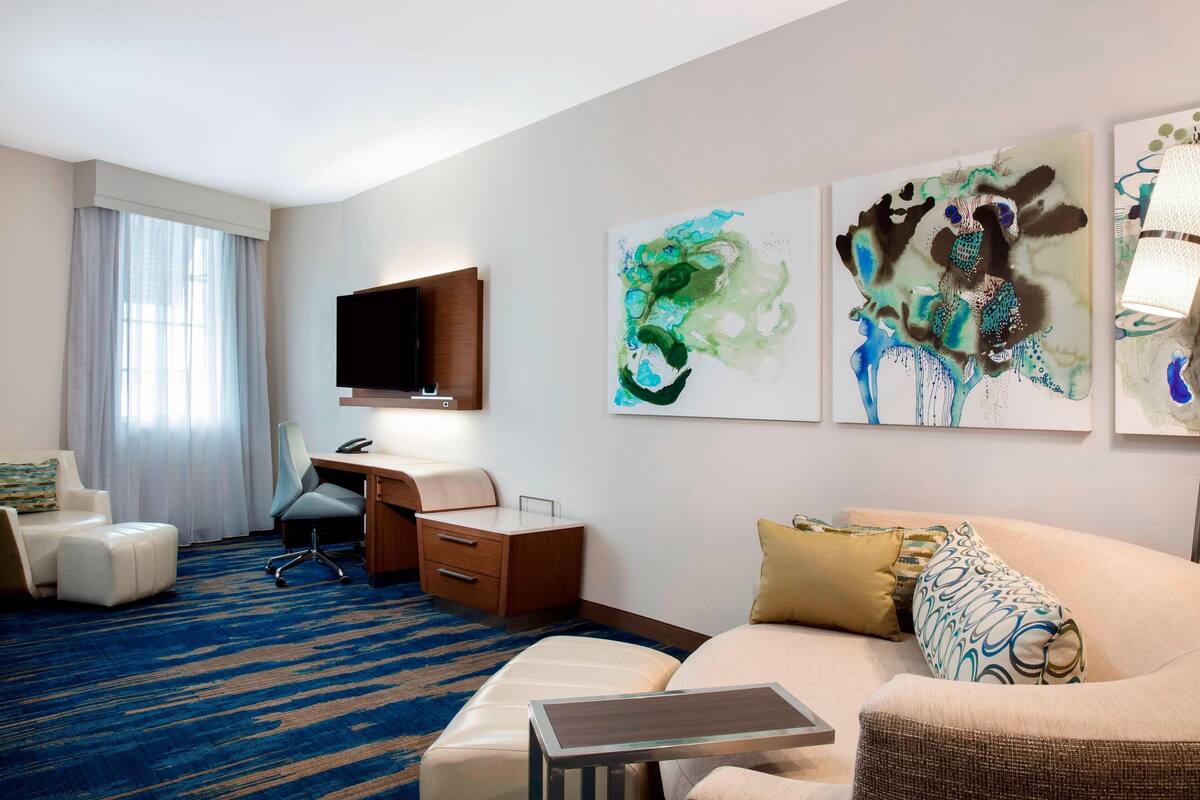 A hotel room with a bed, a desk, a chair, a TV, and artwork on the wall.