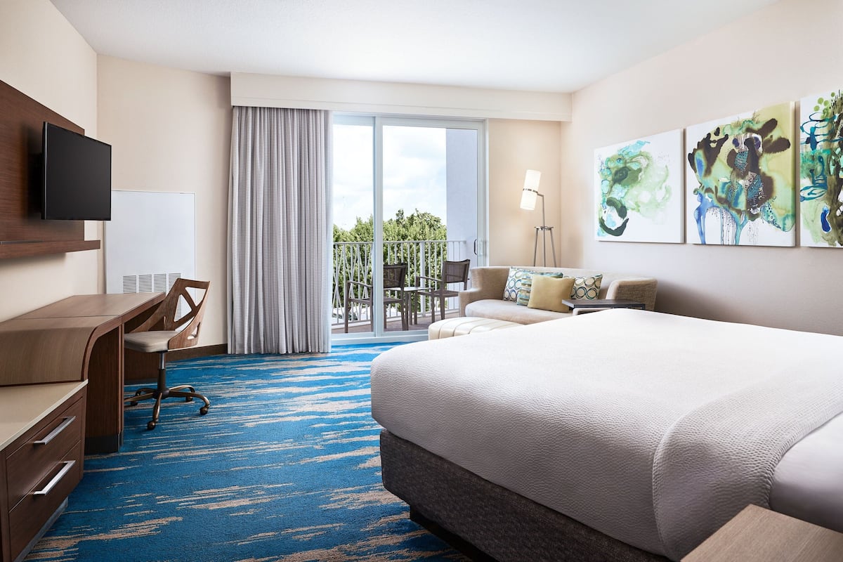 A hotel room with a large bed, a desk, a chair, a flat-screen TV, a large window with curtains, and artwork on the wall.