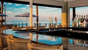 A bar area with a view of the ocean, bar stools, umbrellas, and a variety of bottles.