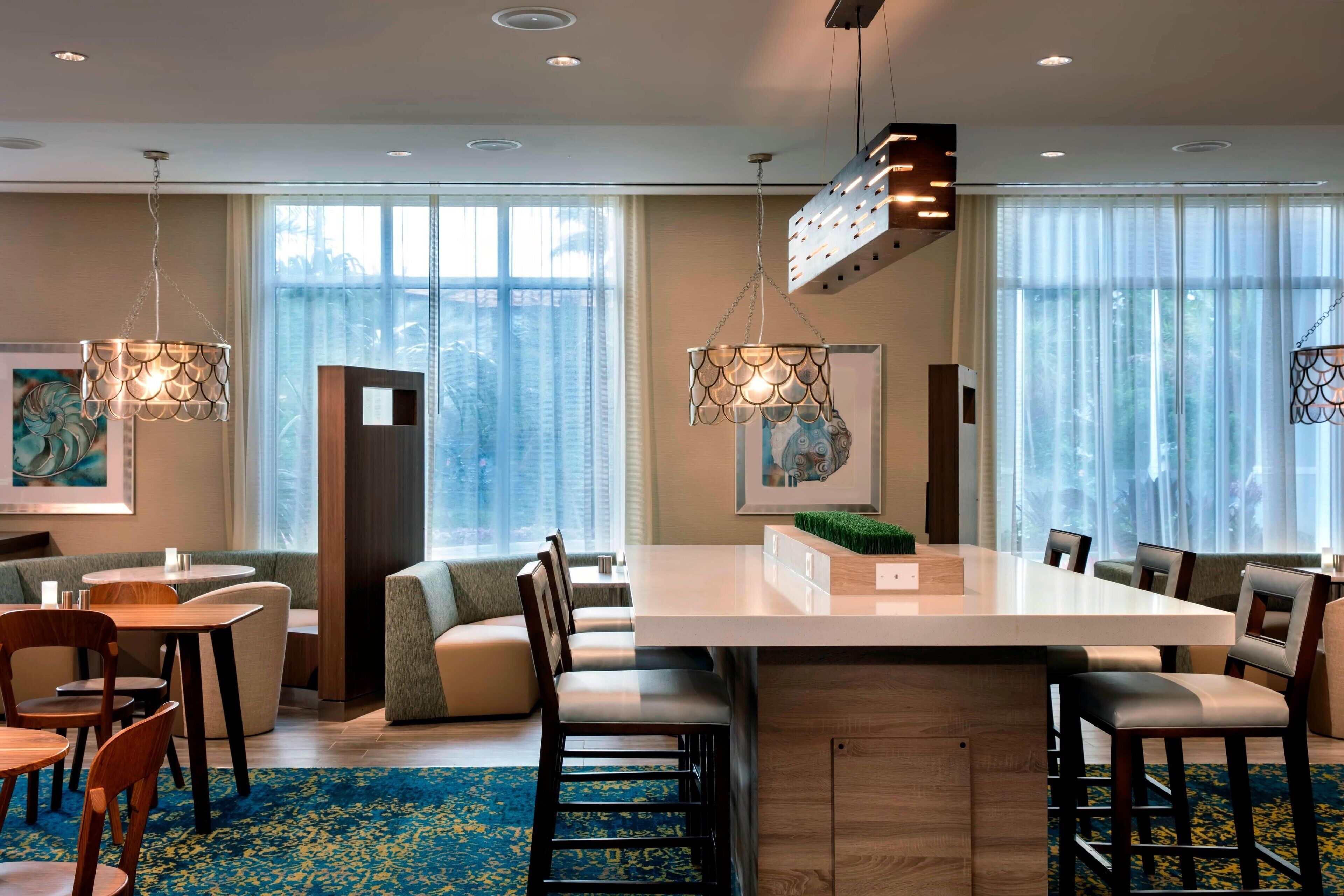 Un comedor moderno de hotel con una gran mesa central, diversas opciones de asiento e iluminación colgante.