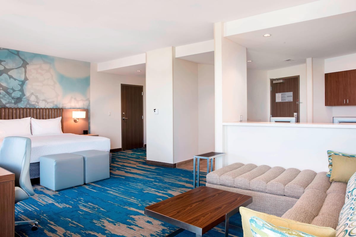 A hotel room with a bed, a sofa, a small table, a desk, and a bathroom visible in the background.