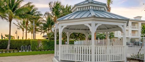 A white gazebo with a metal roof, surrounded by palm trees and a well-maintained lawn.
