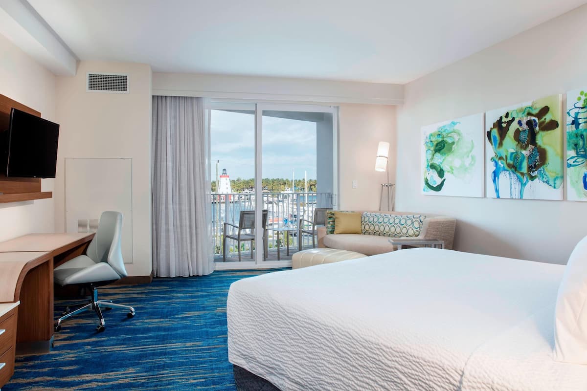 A hotel room with a large bed, a desk, a chair, a sofa, and a balcony with a view of the sea and a lighthouse.