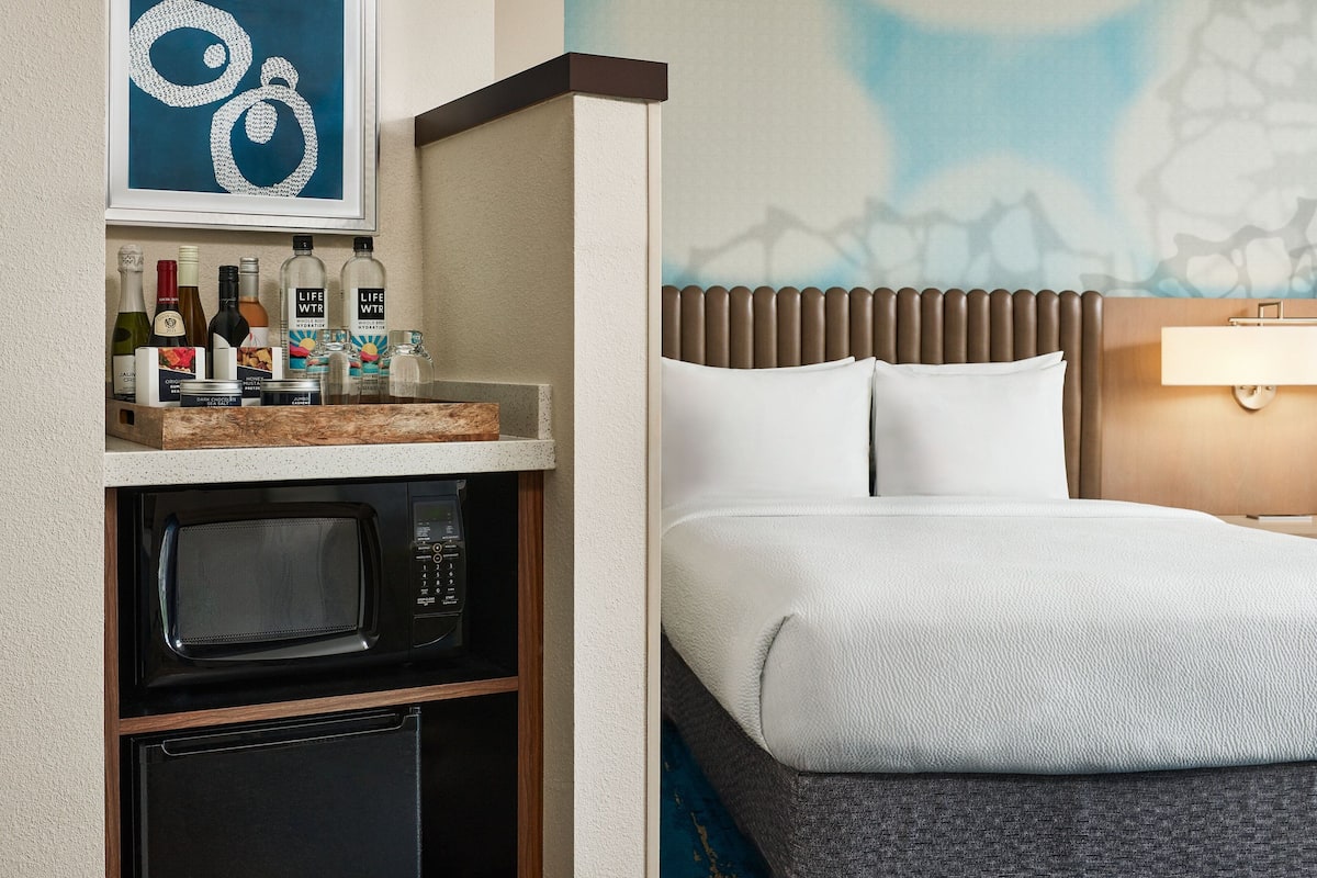 A hotel room with a bed, a microwave, a small refrigerator, a shelf with toiletries, and a framed artwork above the shelf.