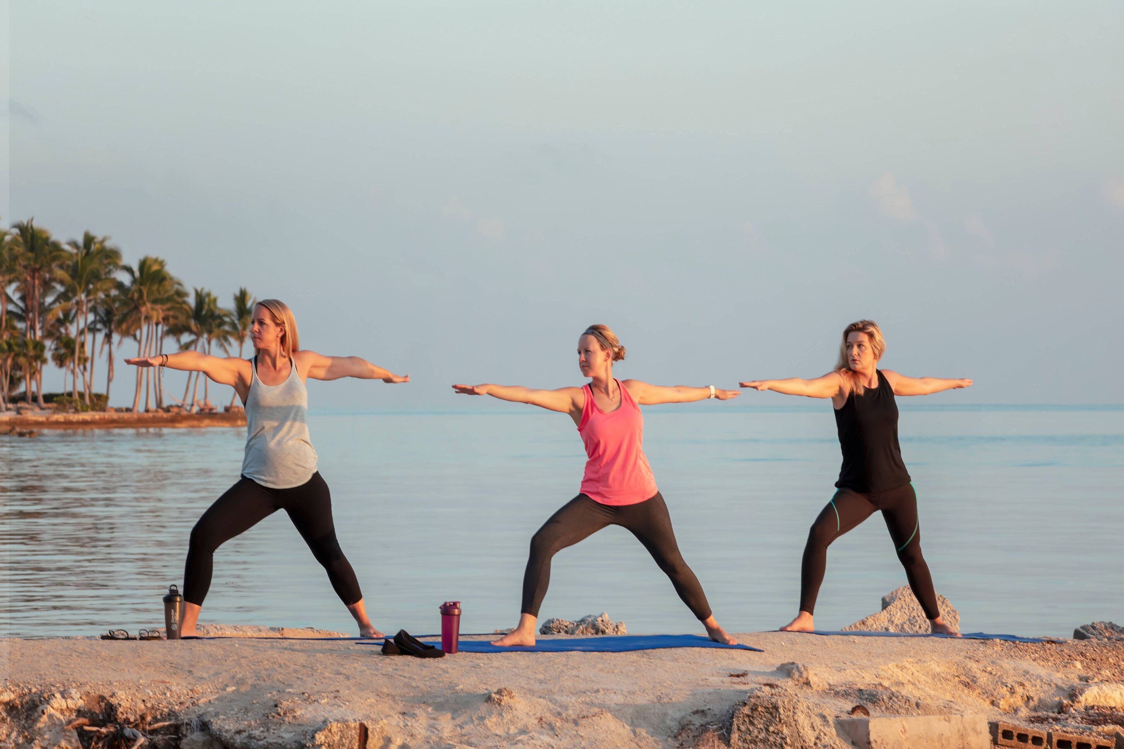 Tres personas practicando yoga en una playa con un mar tranquilo y palmeras al fondo.