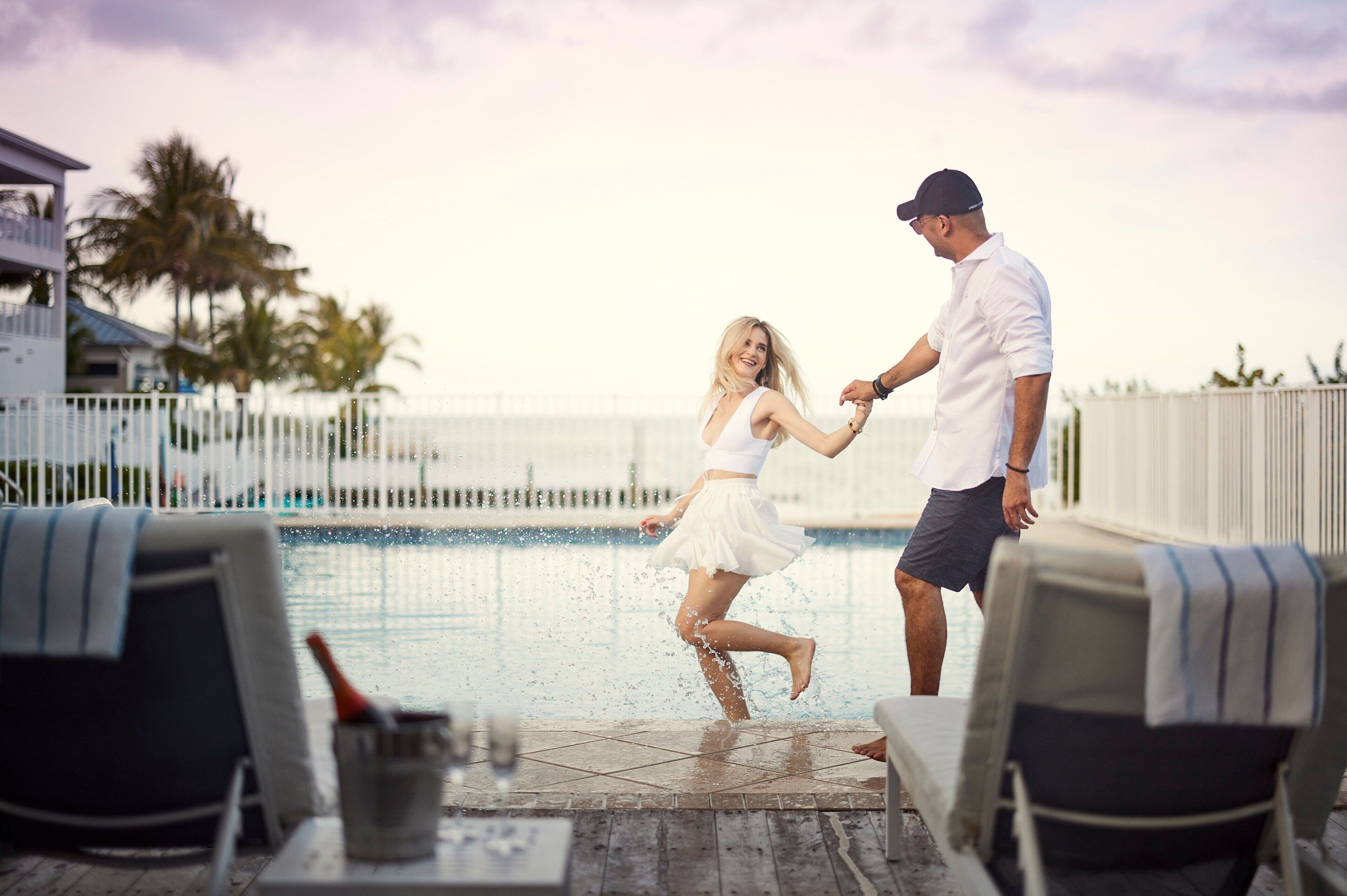 A couple dancing by a poolside with lounge chairs and a fence in the background.
