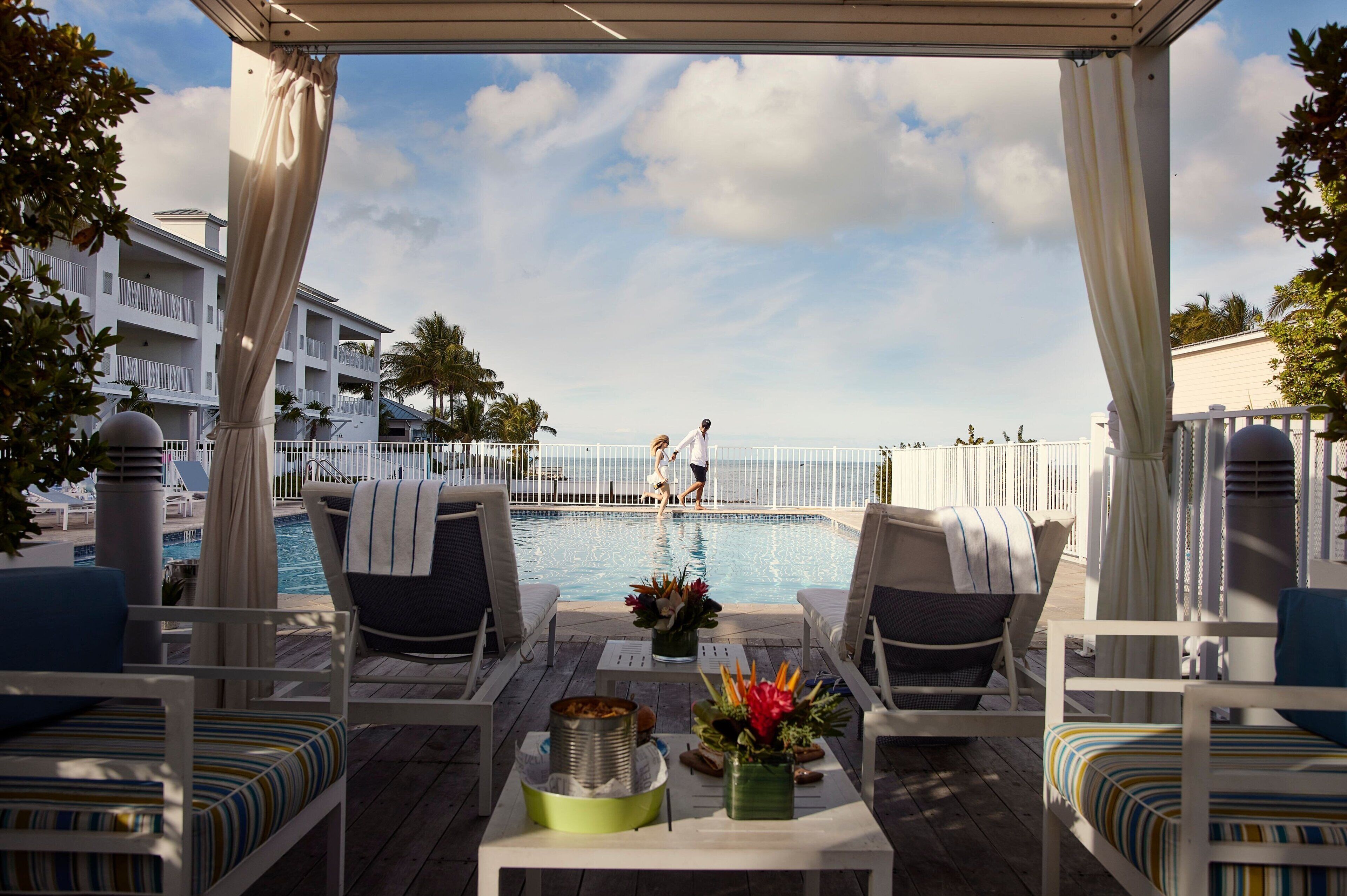A poolside area with lounge chairs, a table with a drink tray, and a view of a pool with people in the background.