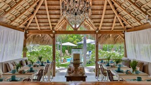 Uma área de refeições ao ar livre com mesas e cadeiras de madeira, um candelabro e vista para uma piscina e vegetação.