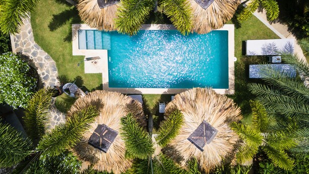 Vista aérea de um resort com uma piscina retangular, cercada por guarda-sóis de palha e vegetação exuberante.