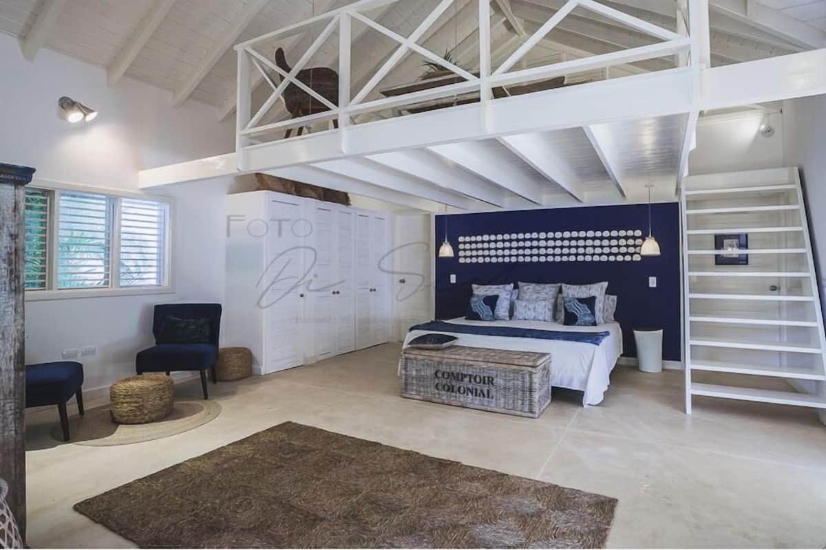 Un dormitorio amplio con zona de altillo, una cama con cabecera estampada, un ventanal grande y una escalera blanca.