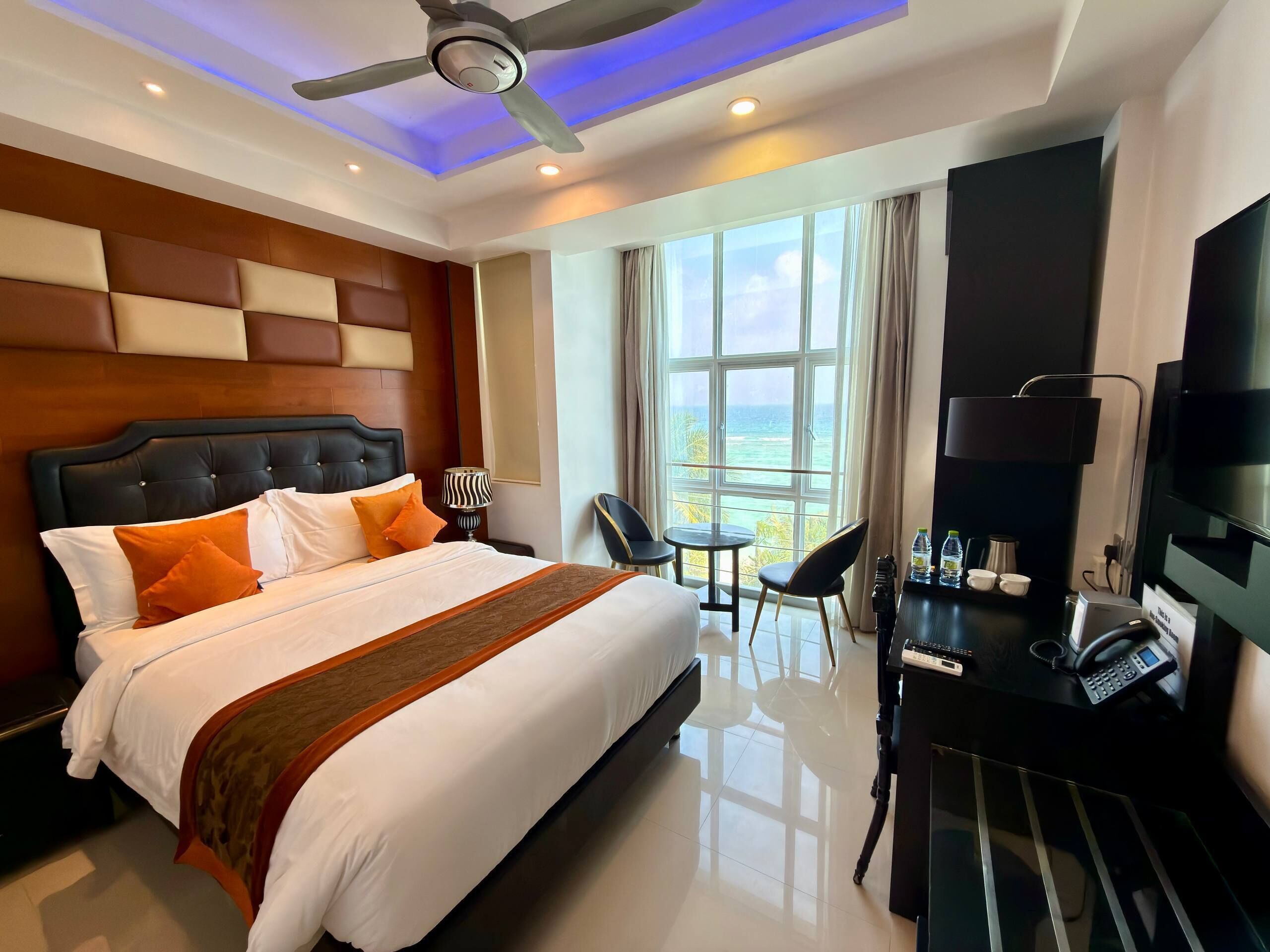 A hotel room with a large bed, a desk with a telephone, and a view of the ocean through large windows.
