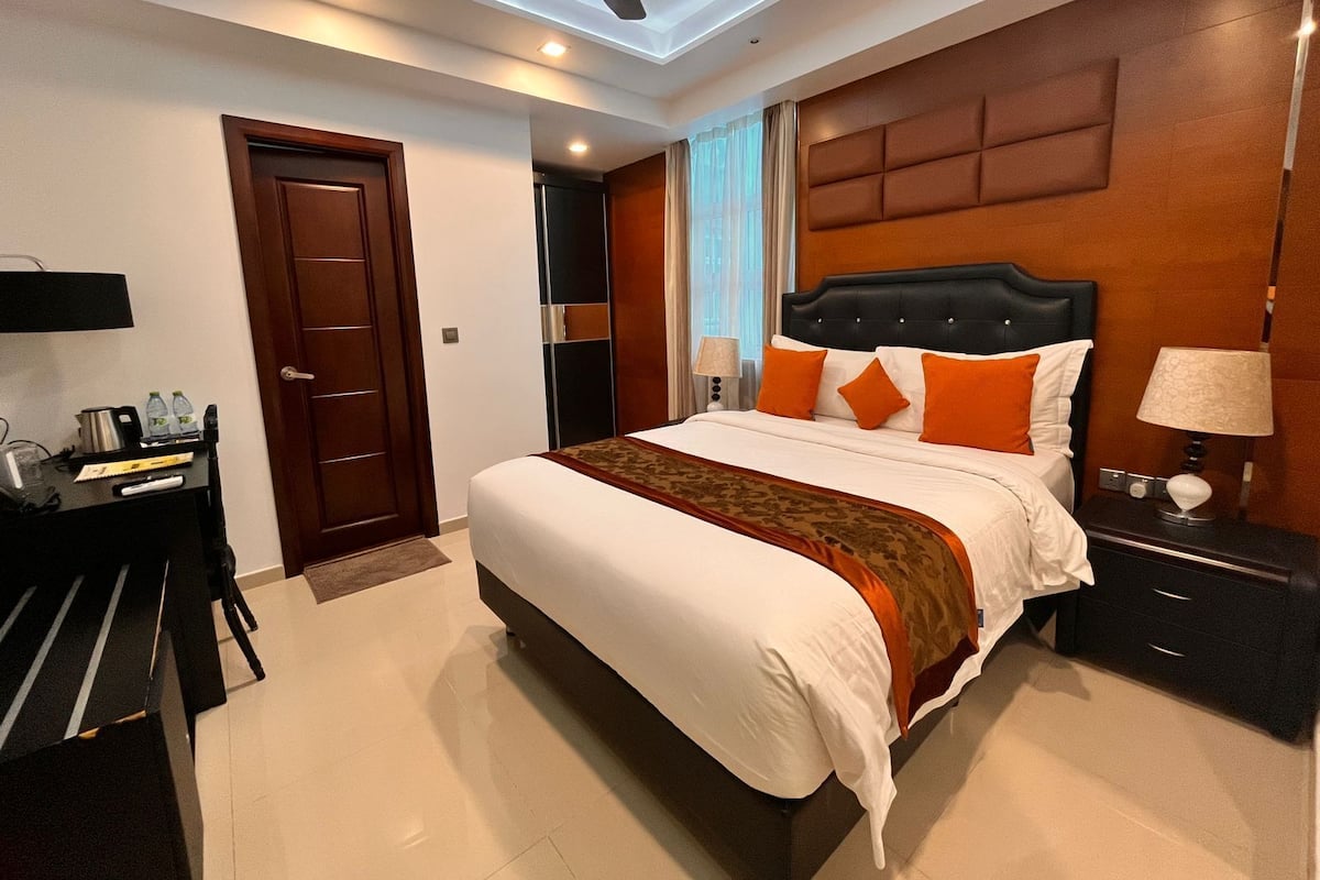 A hotel room with a large bed, a desk with a chair, a nightstand, a ceiling fan, and a window with curtains.