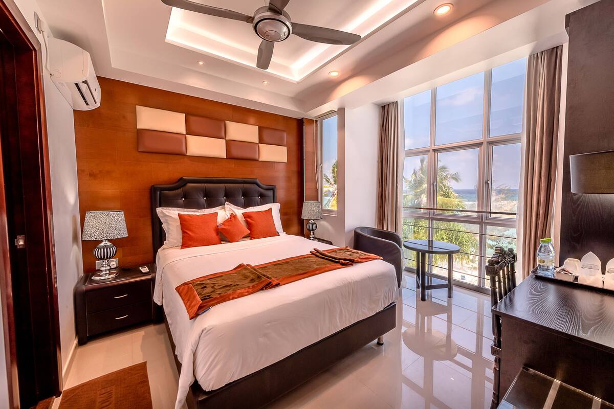 A modern hotel room with a large bed, a ceiling fan, a nightstand with a lamp, a desk with a chair, and a view of the ocean through large windows.