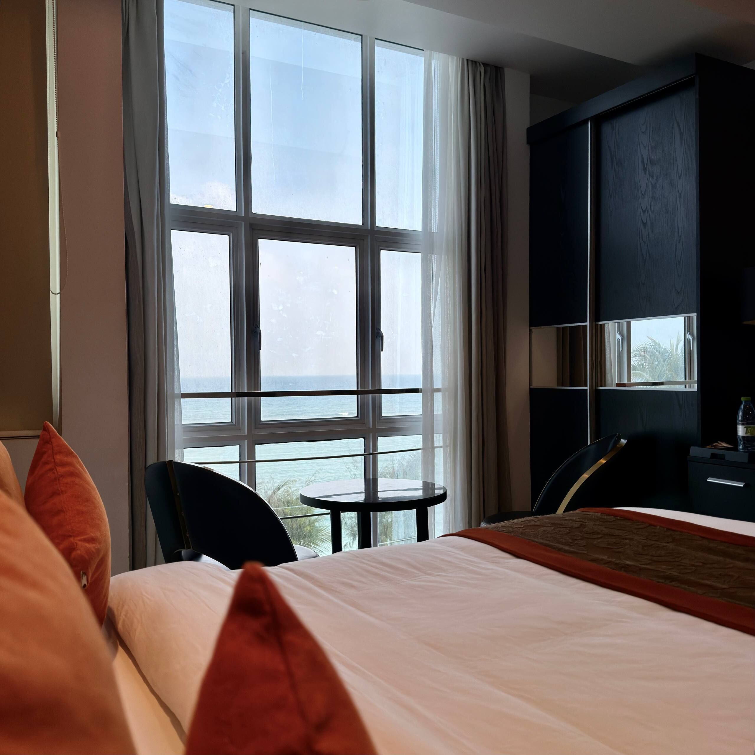 A hotel room with a large window, a bed with white and orange bedding, a small round table, and a dark-colored wardrobe.
