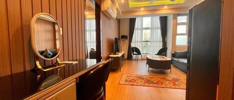 A modern hotel room with a wooden panel wall, a vanity with a mirror, a black leather chair, a sofa, and a ceiling fan.
