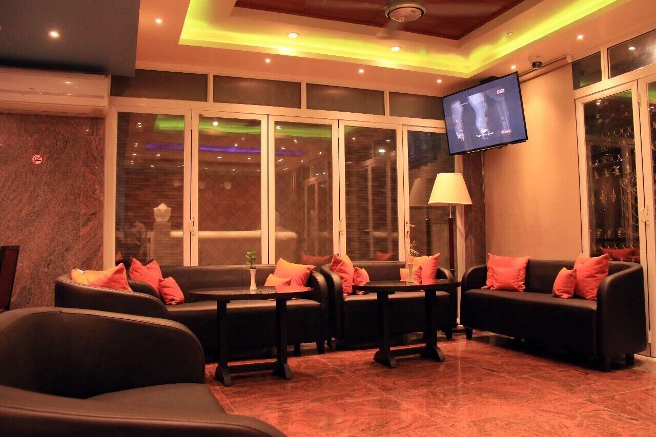 A modern lounge area with black sofas and armchairs, a flat-screen TV mounted on the wall, and a large window with a view of the interior.