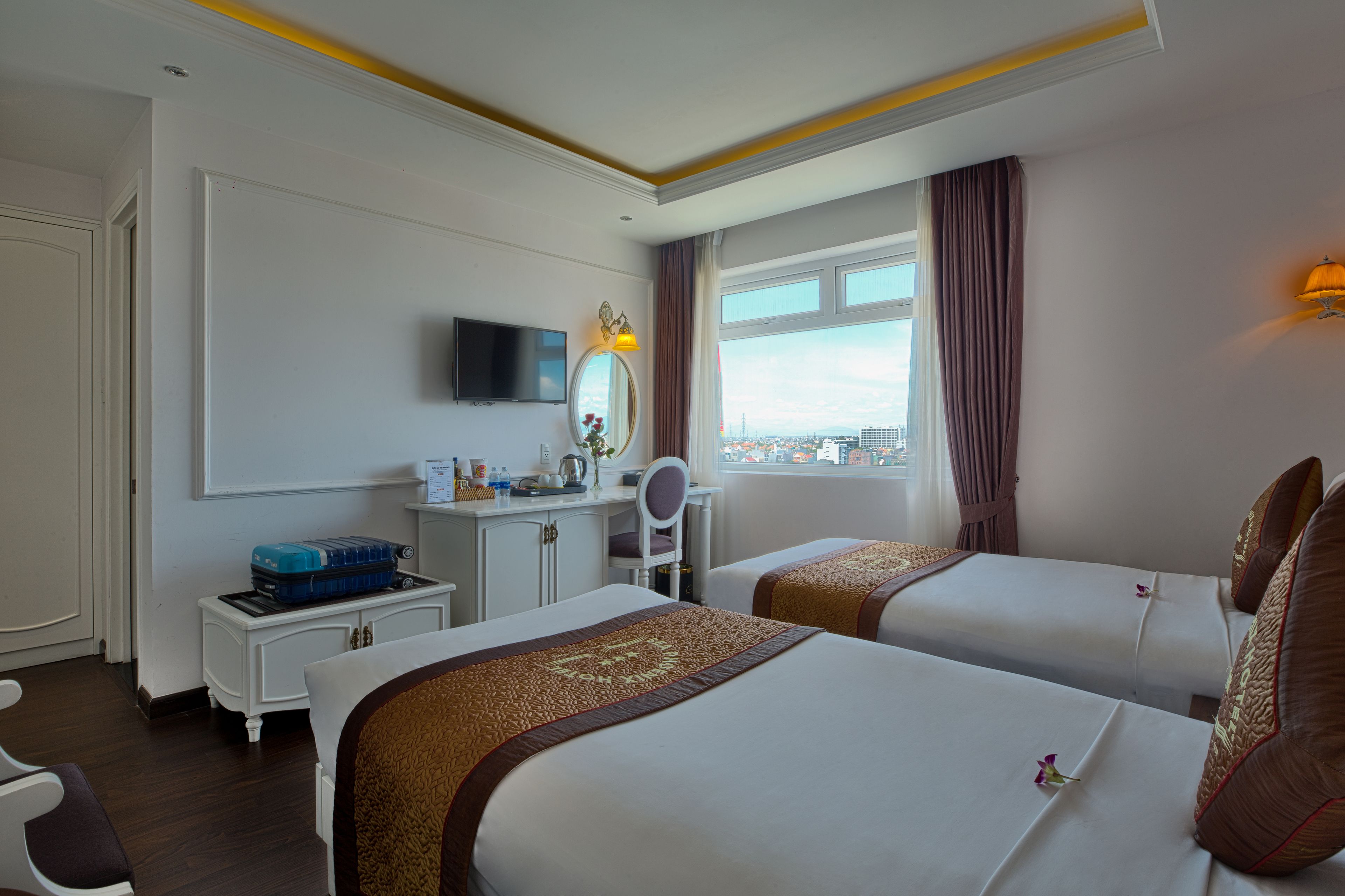 A hotel room with two beds, a desk with a TV, a window with a view, and a cityscape.
