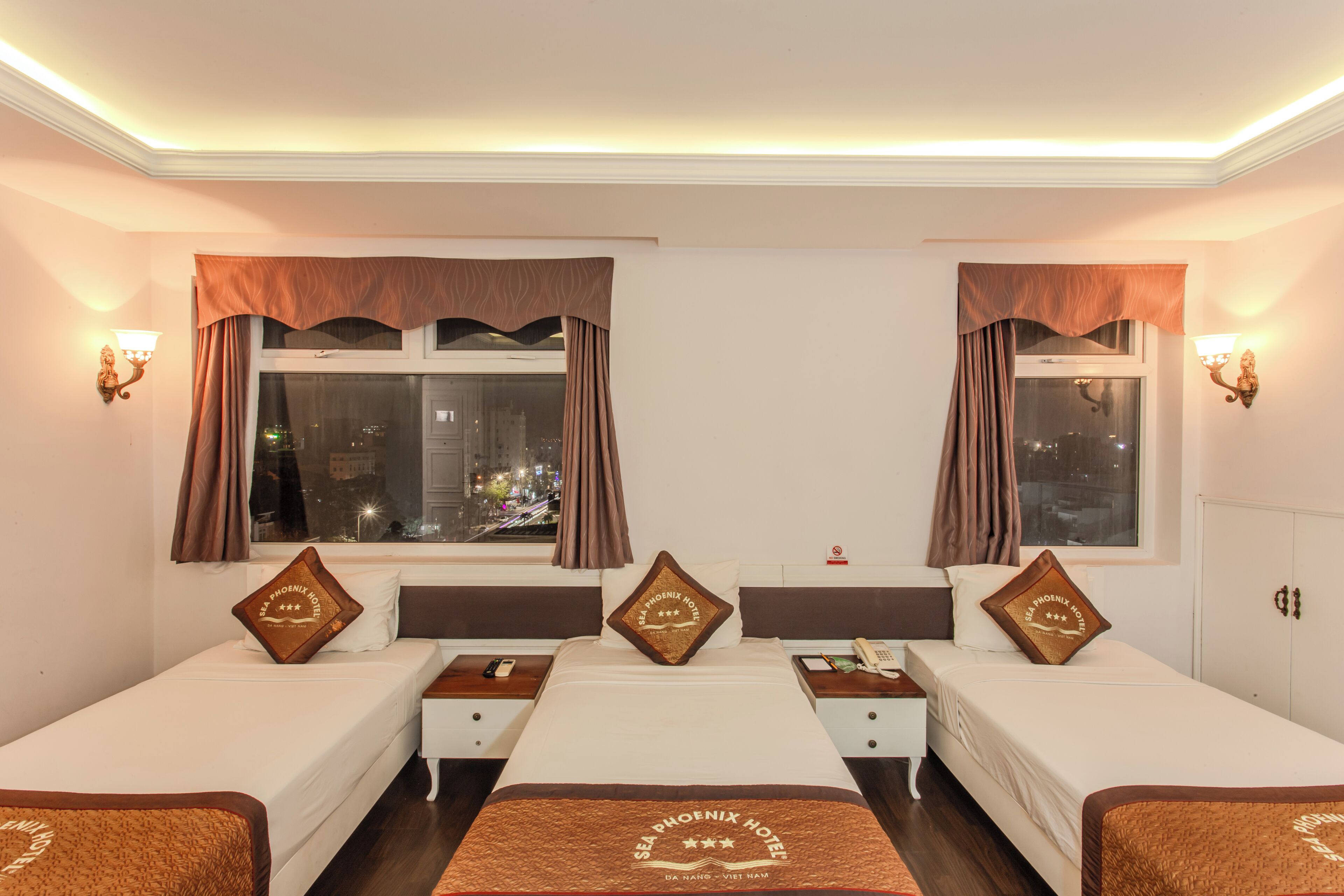 A hotel room with two single beds, a large window with curtains, a city view, and a decorative pillow with the hotel's name.