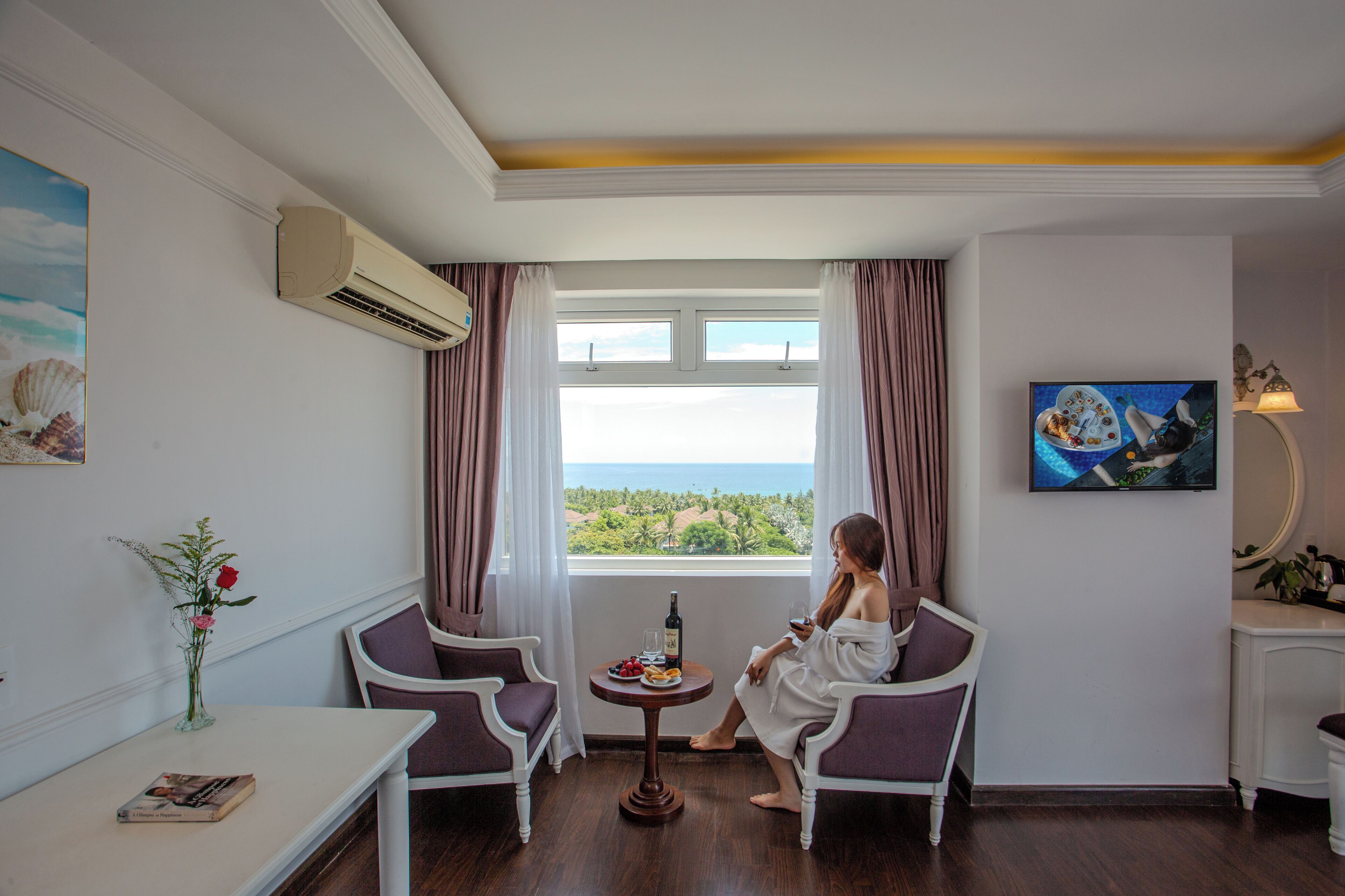 A room with a view of the sea, two chairs, a small table with a bottle and food, and a TV on the wall.