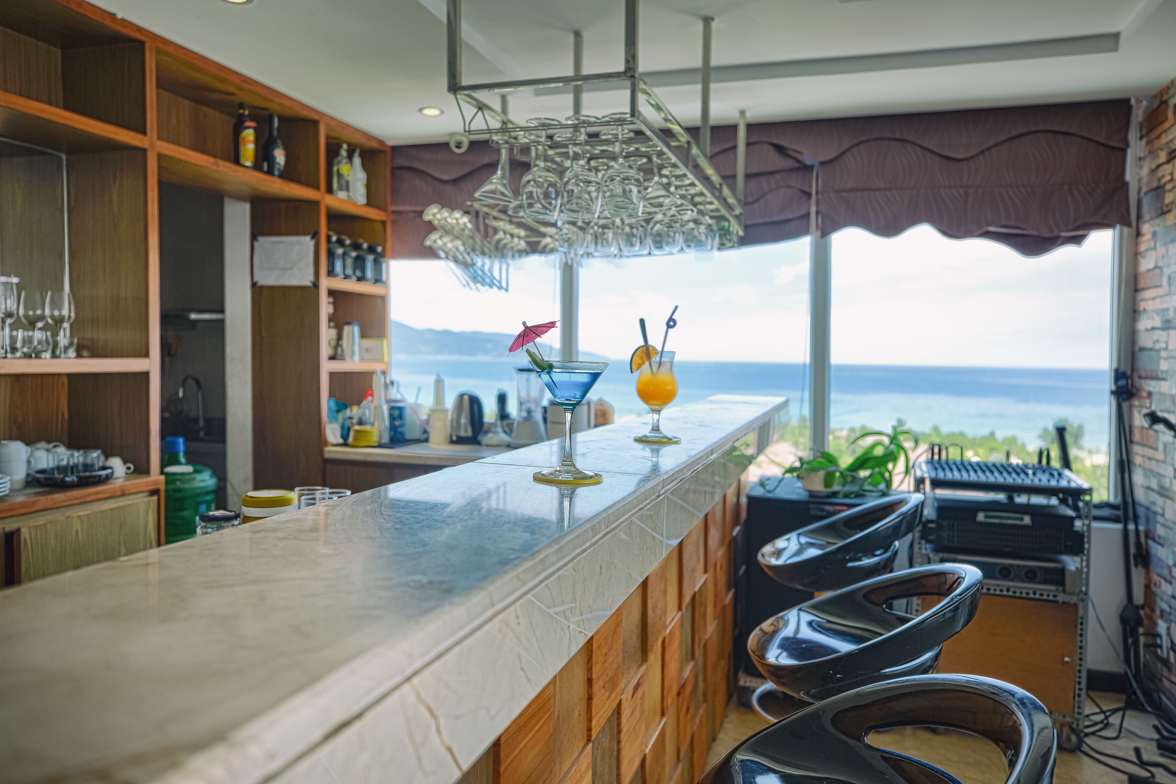 Zona de bar con vista al mar, estanterías con botellas y vasos, y una lámpara colgante.