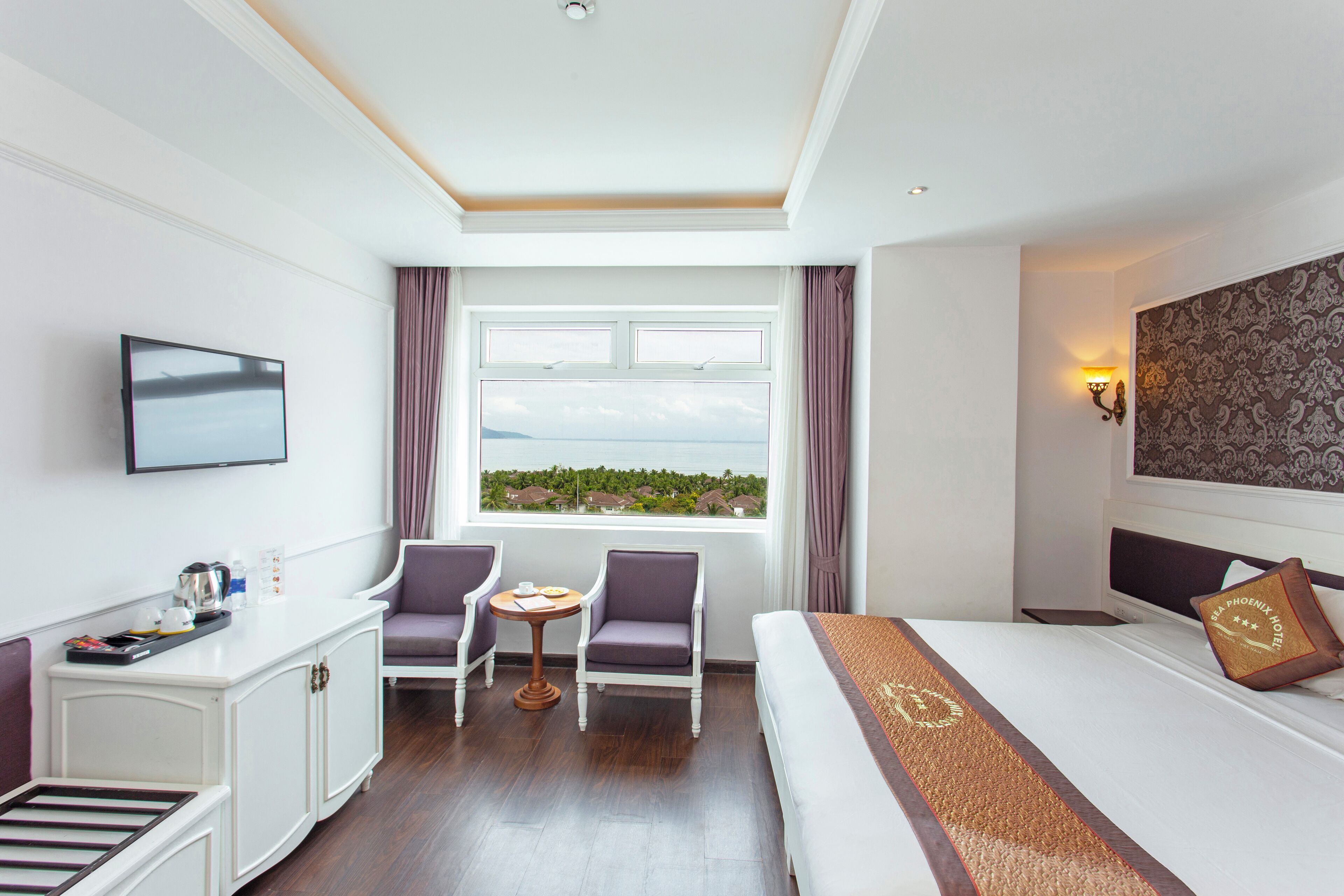 A hotel room with a bed, a desk with a flat-screen TV, two chairs, a small table, and a window with a view of greenery.