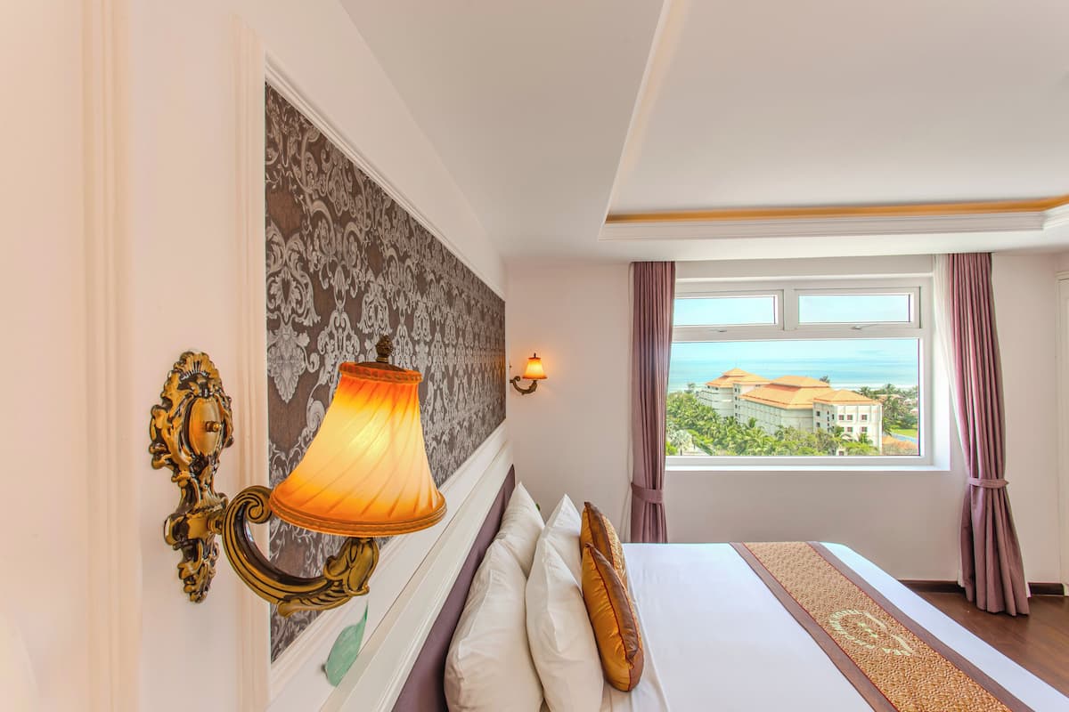 Une chambre d’hôtel avec un lit, une lampe fixée au mur, une vue sur l’océan et une fenêtre avec des rideaux.