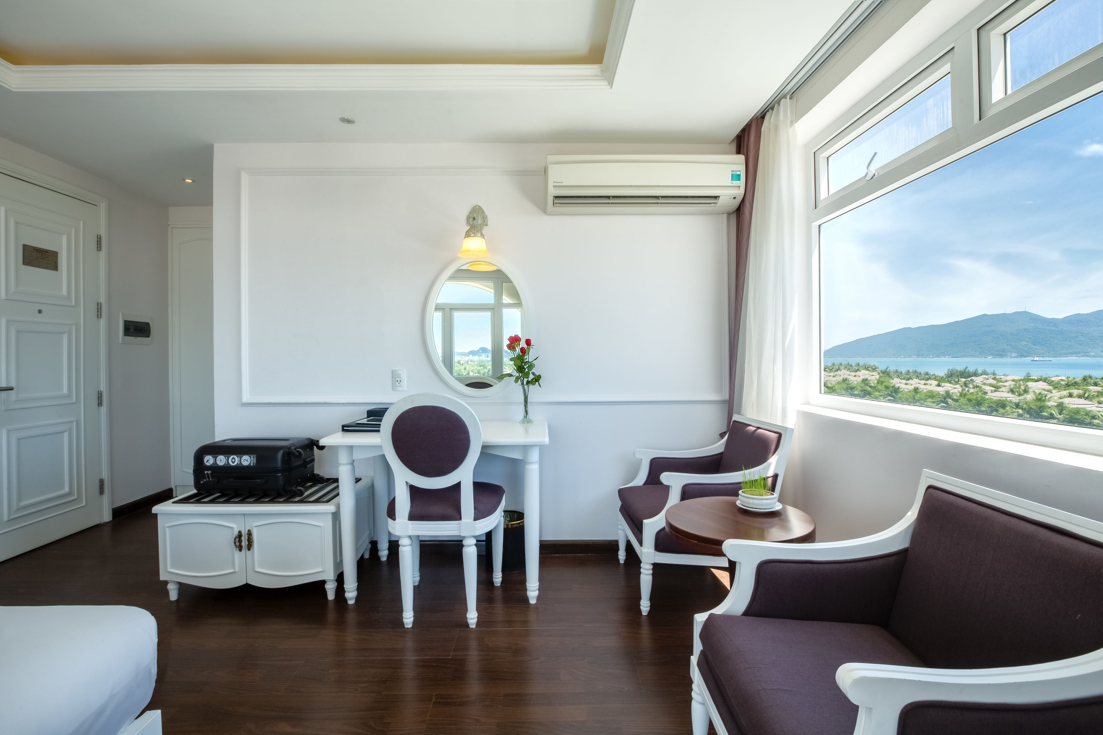 A hotel room with a desk, two chairs, a sofa, and a large window with a view of the sea and mountains.