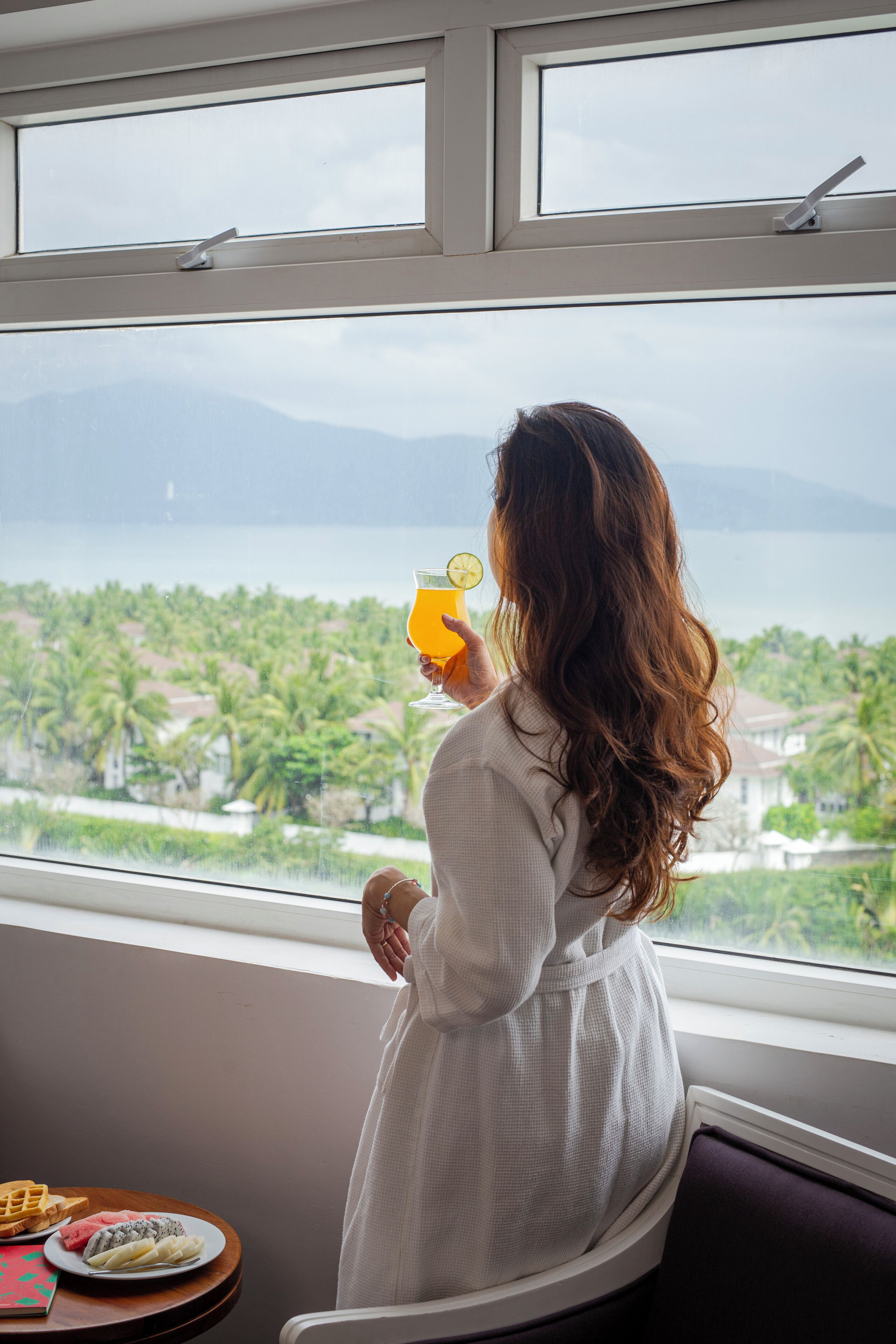 A person in a white dress standing by a window with a view of a coastal landscape.