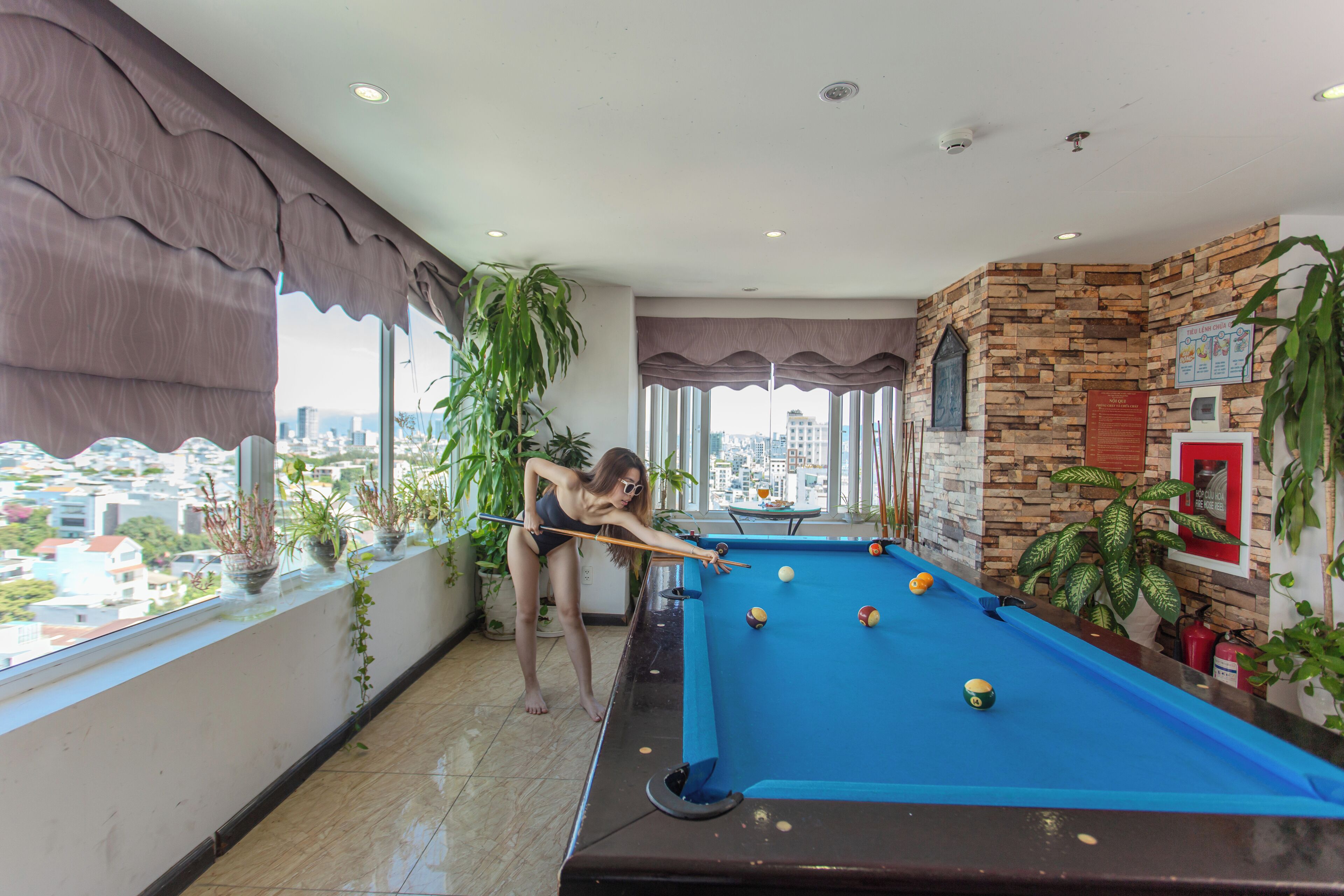 A pool table in a room with large windows, potted plants, and a city view.