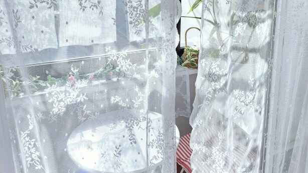 Una habitación con una ventana cubierta por una cortina blanca translúcida con motivos florales.