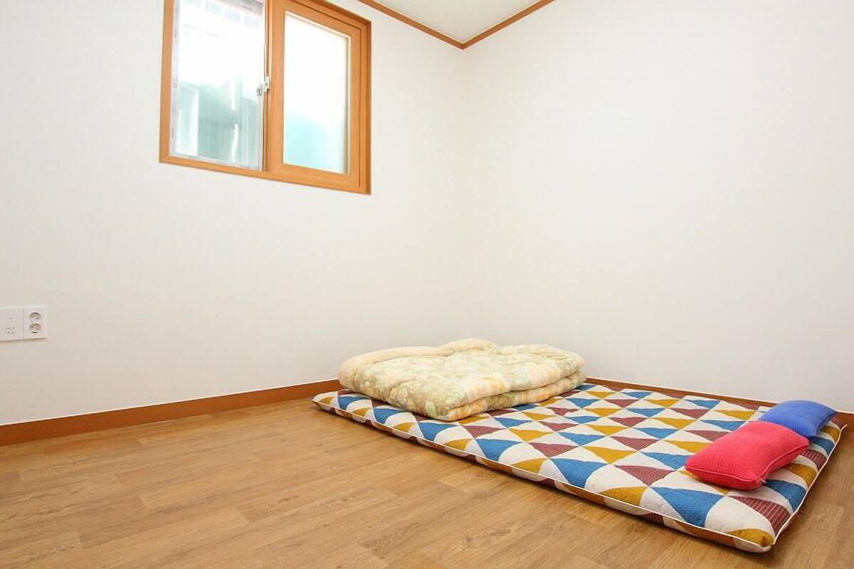 Une petite pièce au mobilier minimaliste, avec un plancher en bois, une fenêtre et un matelas coloré agrémenté de coussins.